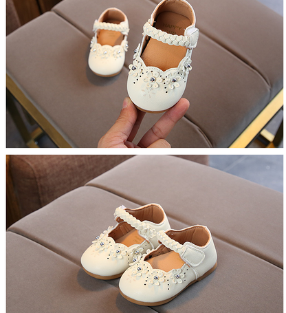 White children's shoes with floral embellishments held by a hand, then placed on a brown surface.
